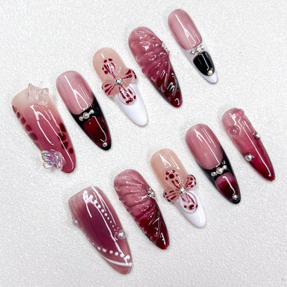 Press-on nails featuring a burgundy-to-black gradient, glossy nude and wine red base tones, 3D butterflies, flower petals, and pearl accents. A luxurious design with gothic elegance and celestial inspiration.