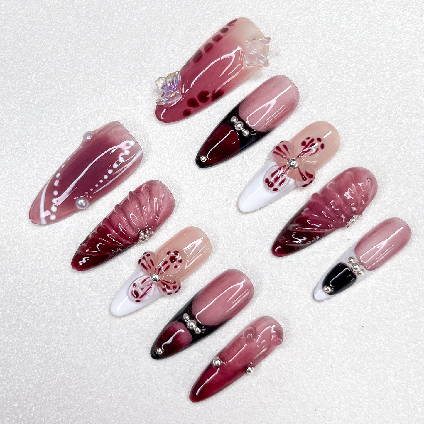 Press-on nails featuring a burgundy-to-black gradient, glossy nude and wine red base tones, 3D butterflies, flower petals, and pearl accents. A luxurious design with gothic elegance and celestial inspiration.