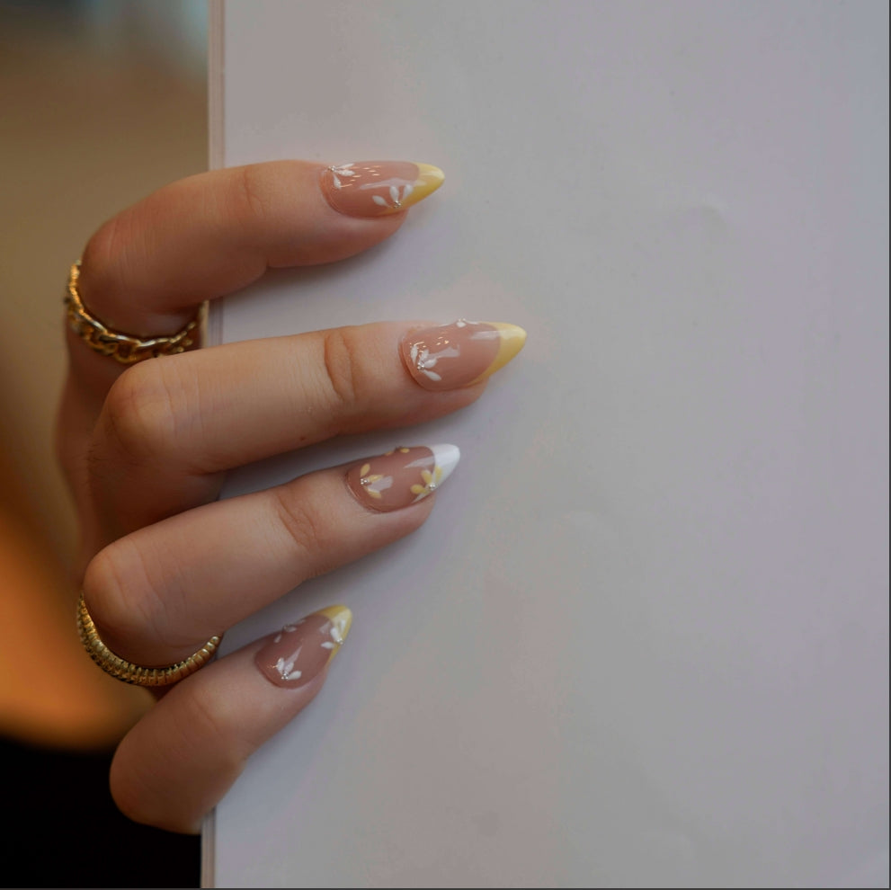 Nude nails with yellow and white French tips, enhanced by delicate daisy-inspired floral accents. Each flower alternates between yellow and white petals with a shimmering diamond at the center, evoking the tranquil beauty of a sunlit meadow.