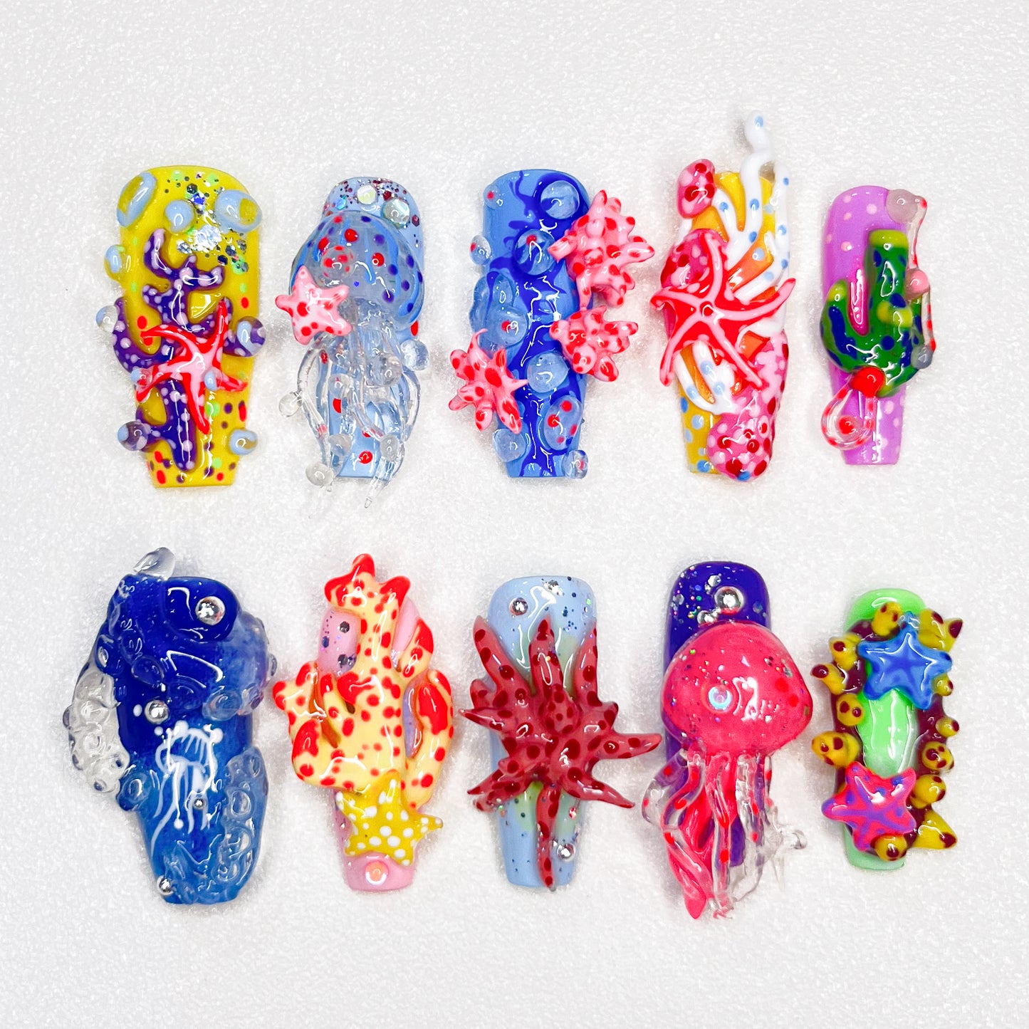 A set of ocean-themed press-on nails showcasing intricate 3D details of coral reefs, starfish, jellyfish, and vibrant underwater designs in blue, yellow, red, and green tones.