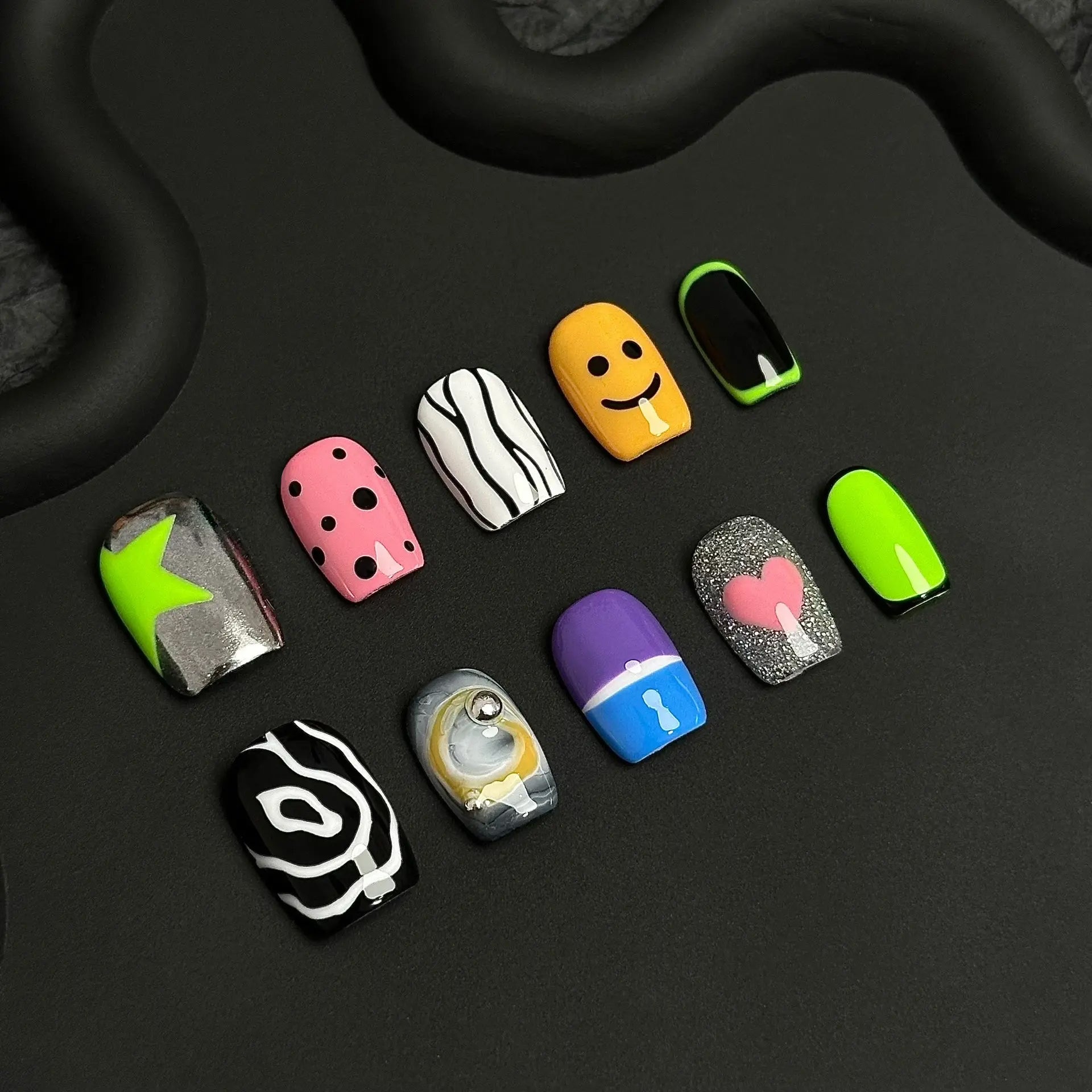 A press-on nail set featuring neon green, pink polka dots, smiley faces, abstract black-and-white patterns, and glitter accents, creating a playful and pop-art-inspired design.