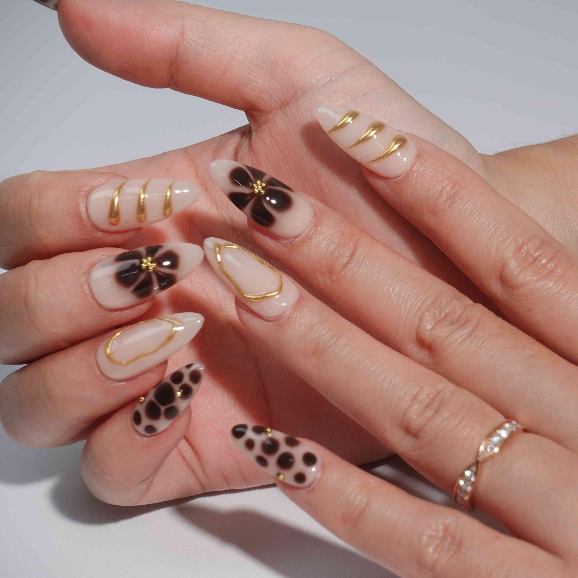 A stunning set of handmade press-on nails featuring bold brown floral prints, delicate gold embellishments, and intricate designs on a nude beige background, creating a refined yet artistic look.