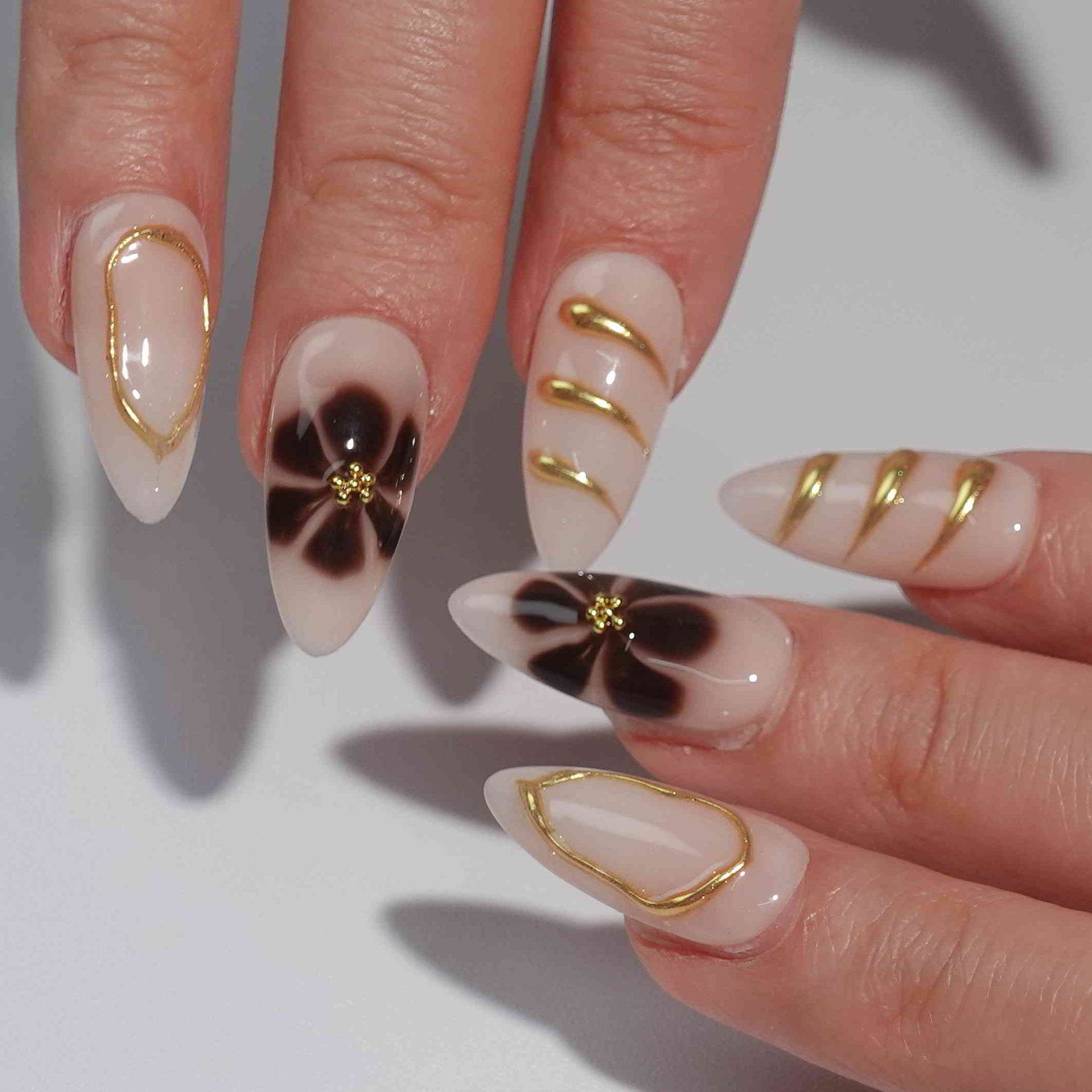 A stunning set of handmade press-on nails featuring bold brown floral prints, delicate gold embellishments, and intricate designs on a nude beige background, creating a refined yet artistic look.