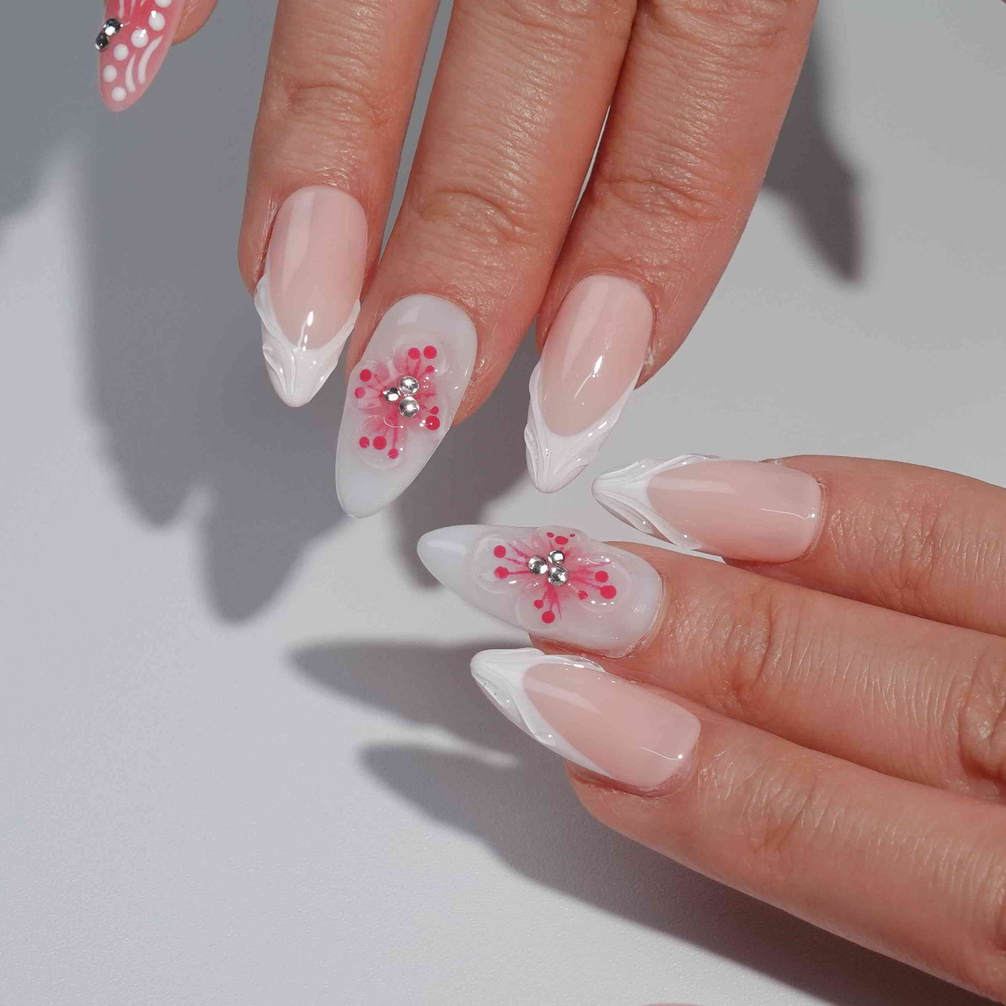 A handmade set of press-on nails showcasing pink gradients, intricate white petal-like designs, and 3D floral centers adorned with rhinestones, evoking the charm of blooming spring flowers under soft sunlight.