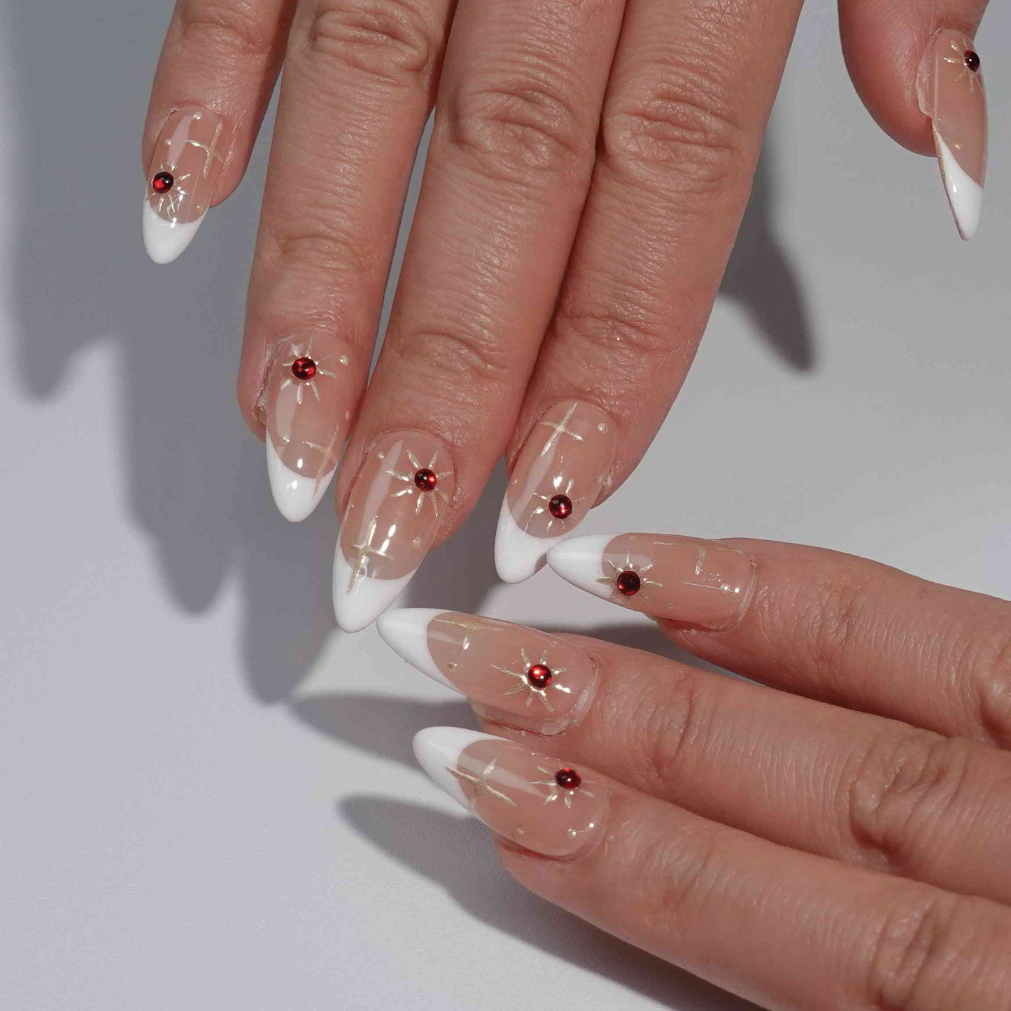 Press-on nails with a nude base, white French tips, decorated with gold star patterns and striking red gem embellishments, creating an elegant and festive design.