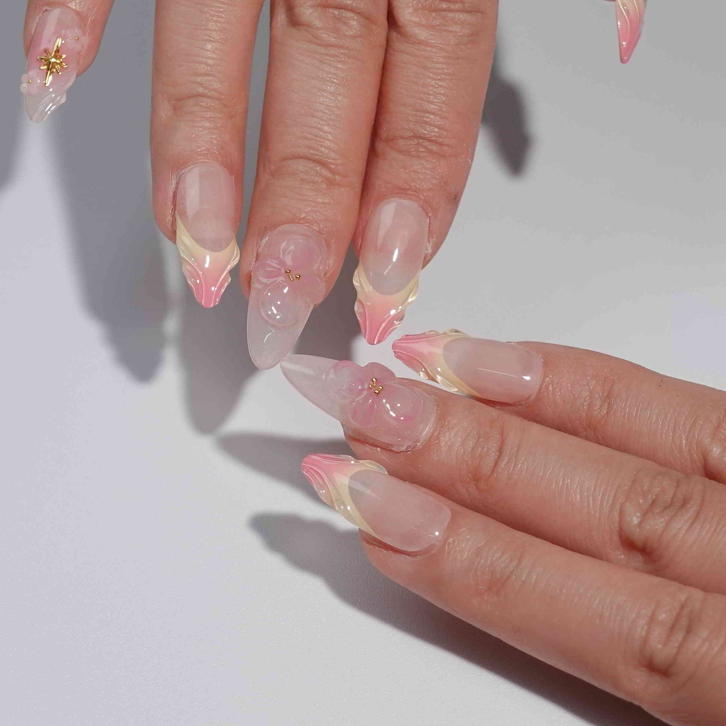 A handmade set of press-on nails showcasing pink and peach ombré designs with sculpted petal-like tips, complemented by 3D floral accents and golden embellishments, exuding elegance and delicate charm.