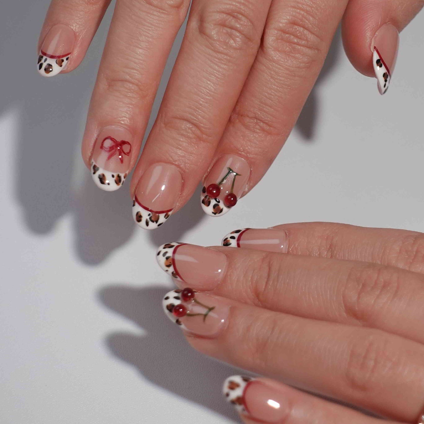 A set of press-on nails with a nude pink base and intricate details. The designs include leopard print French tips, red cherry accents with green stems, and delicate red bows. This artistic nail set blends wild animal motifs with soft, playful touches for a unique fashion statement.