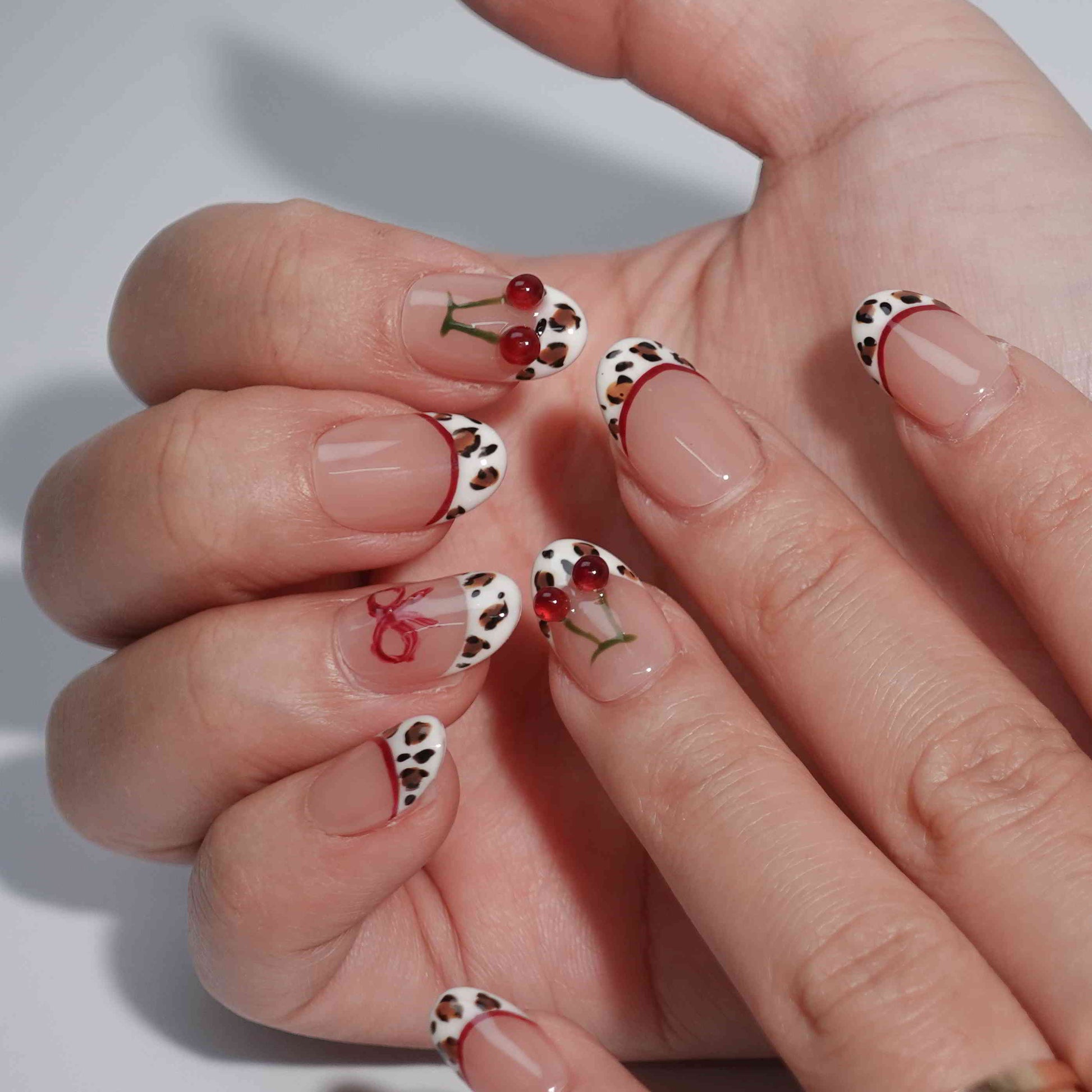 A set of press-on nails with a nude pink base and intricate details. The designs include leopard print French tips, red cherry accents with green stems, and delicate red bows. This artistic nail set blends wild animal motifs with soft, playful touches for a unique fashion statement.