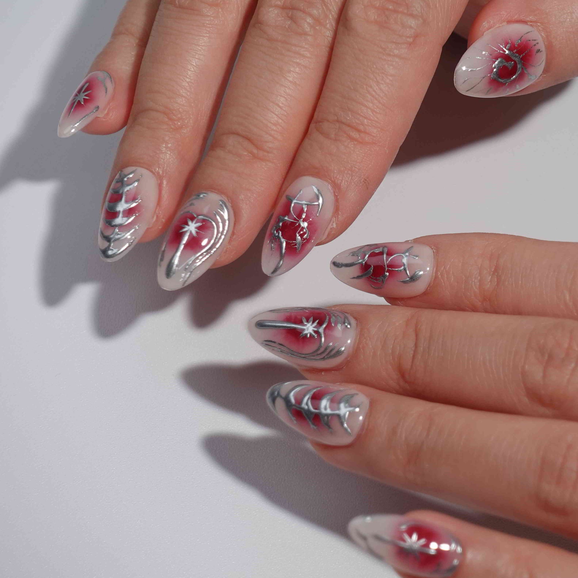 A striking press-on nail set with a red gradient background, adorned with silver metallic details, including abstract floral designs, delicate vines, and celestial star motifs. A perfect choice for a dramatic yet sophisticated look.