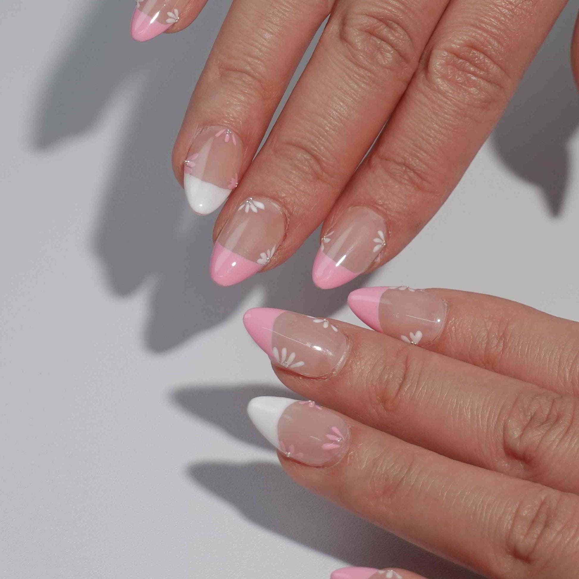 A feminine nail design named "Blush Petals," showcasing pink and white French tips with soft floral accents in matching hues. Each flower is delicately painted with a shimmering rhinestone center, evoking the romance of spring gardens in full bloom.