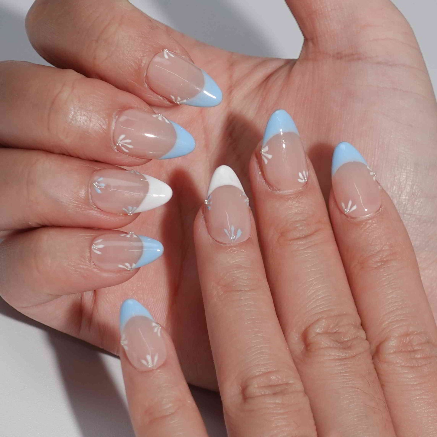 Nude nails featuring blue and white French tips with elegant flower patterns. The petals alternate in blue and white, complemented by a subtle diamond at the center, offering a refined and breezy aesthetic.