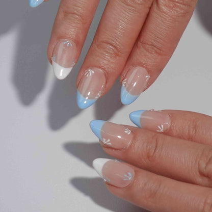 Nude nails featuring blue and white French tips with elegant flower patterns. The petals alternate in blue and white, complemented by a subtle diamond at the center, offering a refined and breezy aesthetic.