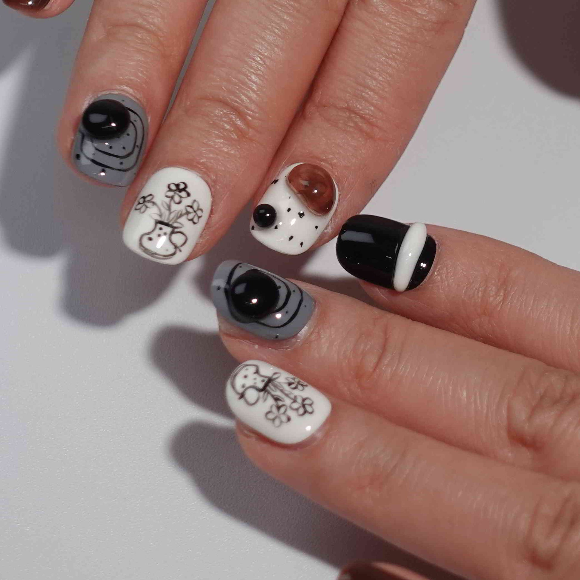 A set of press - on nails with a sophisticated mix of black, white, and brown colors, featuring elegant floral and geometric designs, neatly arranged in a transparent case. The nails have an artistic and stylish finish, ideal for both casual and formal events.