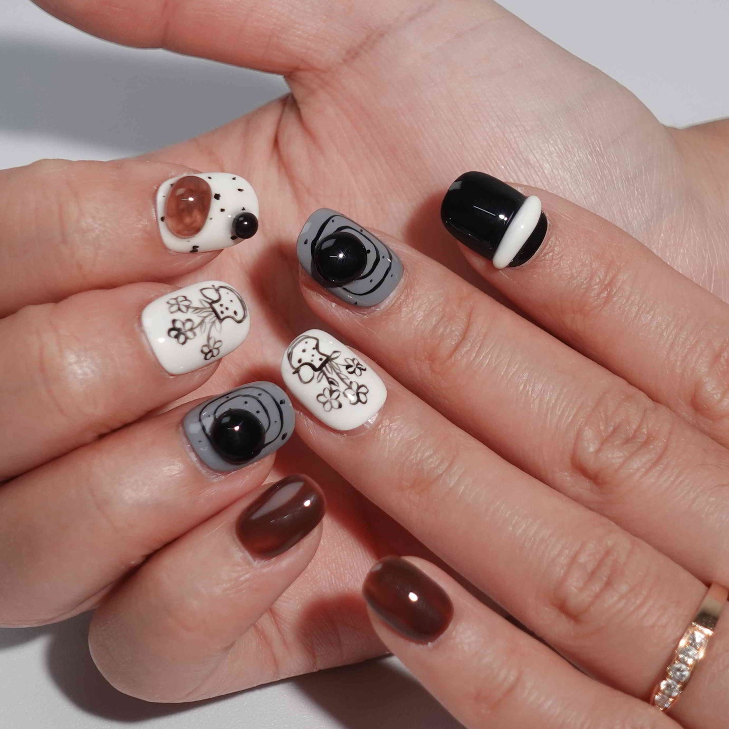 A set of press - on nails with a sophisticated mix of black, white, and brown colors, featuring elegant floral and geometric designs, neatly arranged in a transparent case. The nails have an artistic and stylish finish, ideal for both casual and formal events.