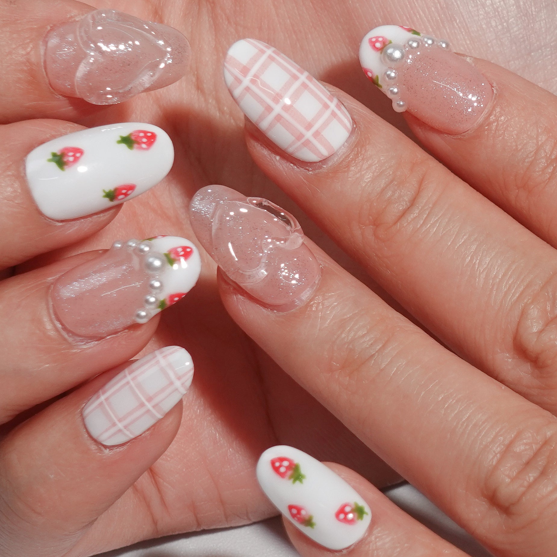 A fruity and feminine press-on nail design featuring pink glitter, plaid patterns, and tiny red strawberries with green leaves. Some nails are adorned with pearls and a 3D bow, creating a playful yet elegant look.