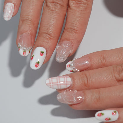 A fruity and feminine press-on nail design featuring pink glitter, plaid patterns, and tiny red strawberries with green leaves. Some nails are adorned with pearls and a 3D bow, creating a playful yet elegant look.