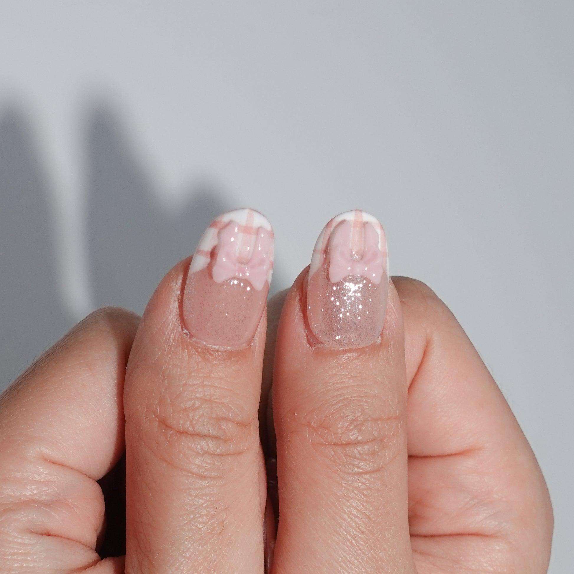 A fruity and feminine press-on nail design featuring pink glitter, plaid patterns, and tiny red strawberries with green leaves. Some nails are adorned with pearls and a 3D bow, creating a playful yet elegant look.