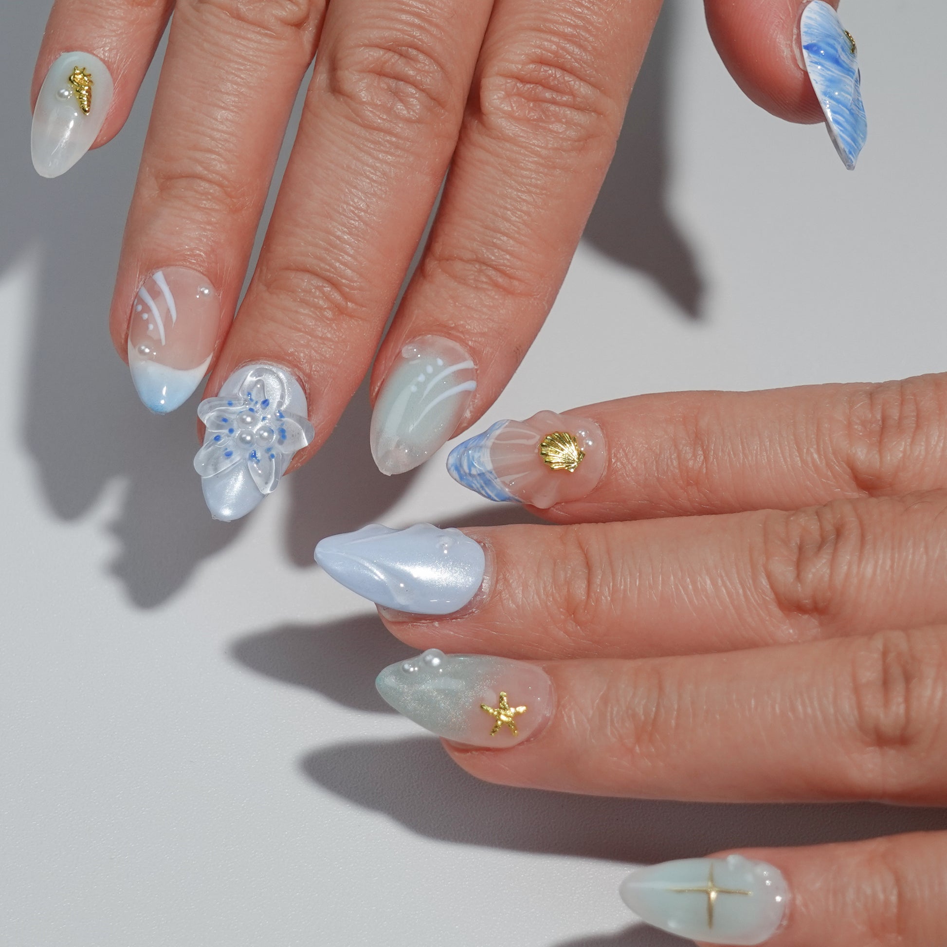 A set of handmade press-on nails showcasing soft blue and pastel gradients, detailed flower designs, starfish motifs, and pearl embellishments. These nails radiate an ocean-inspired elegance, perfect for a sophisticated and serene look.