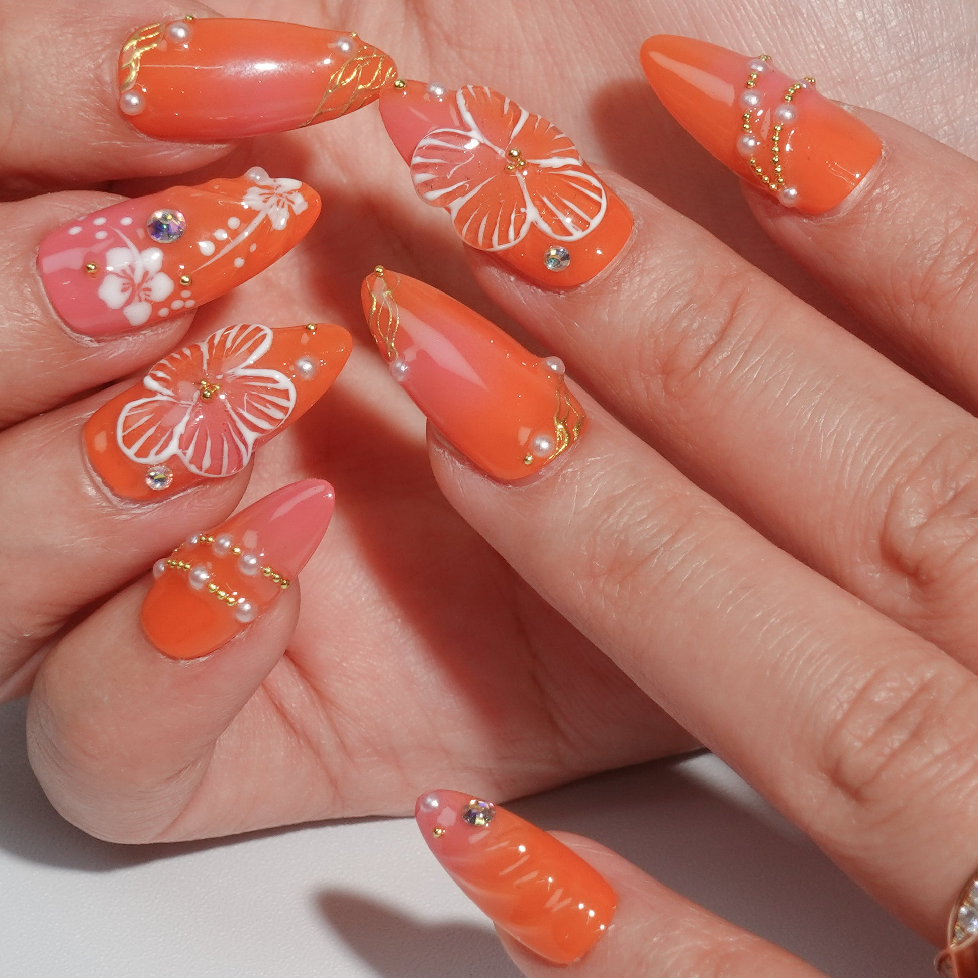 A vibrant coral press-on nail set featuring hand-painted white hibiscus flowers, sparkling rhinestones, and golden accents, evoking the warmth and beauty of a tropical getaway. Perfect for summer-inspired looks.