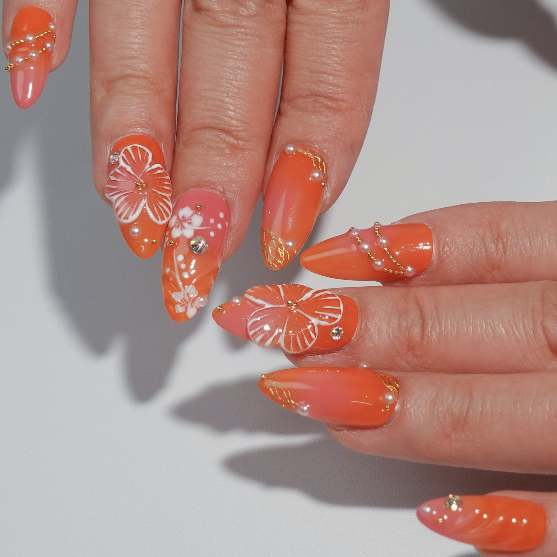 A vibrant coral press-on nail set featuring hand-painted white hibiscus flowers, sparkling rhinestones, and golden accents, evoking the warmth and beauty of a tropical getaway. Perfect for summer-inspired looks.