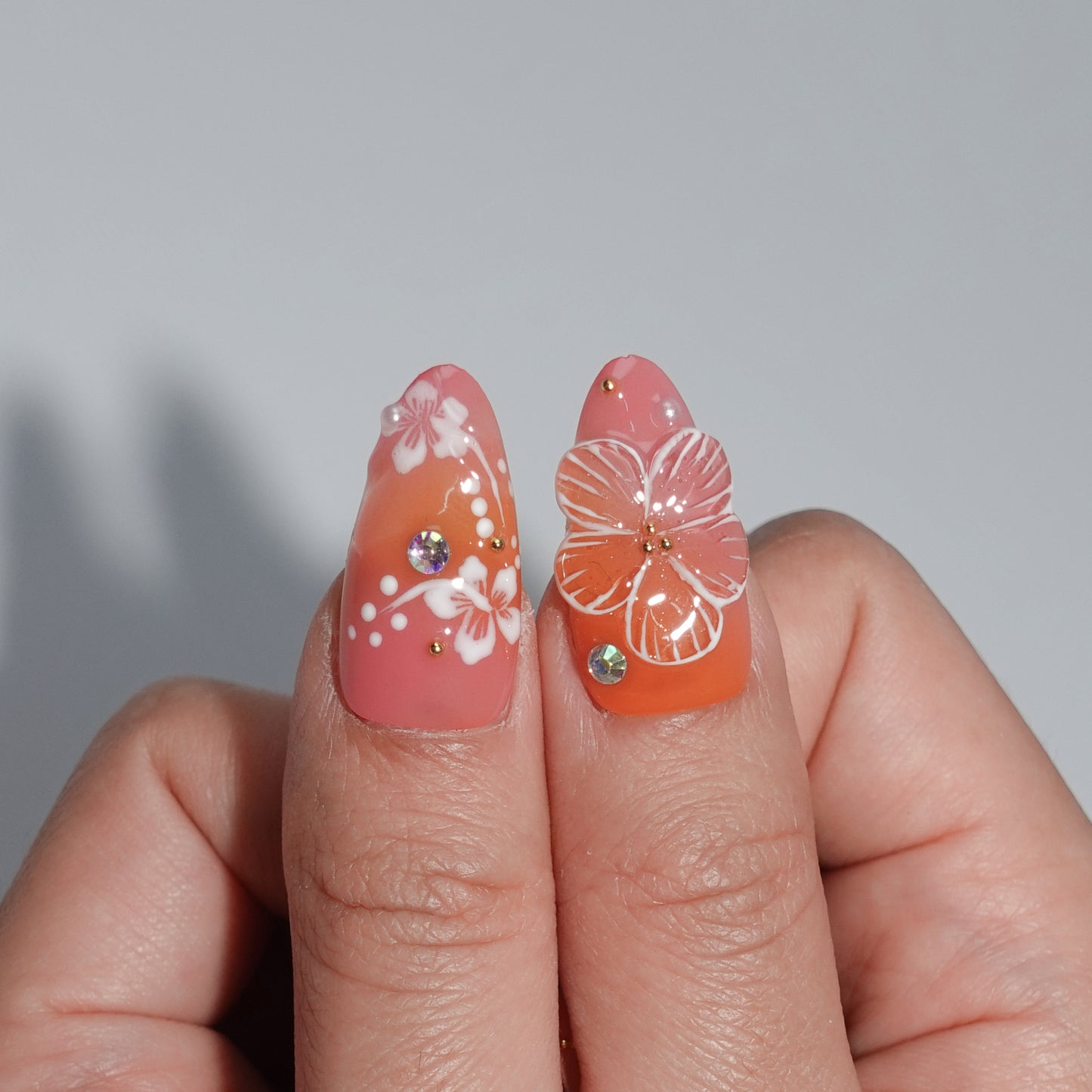 A vibrant coral press-on nail set featuring hand-painted white hibiscus flowers, sparkling rhinestones, and golden accents, evoking the warmth and beauty of a tropical getaway. Perfect for summer-inspired looks.