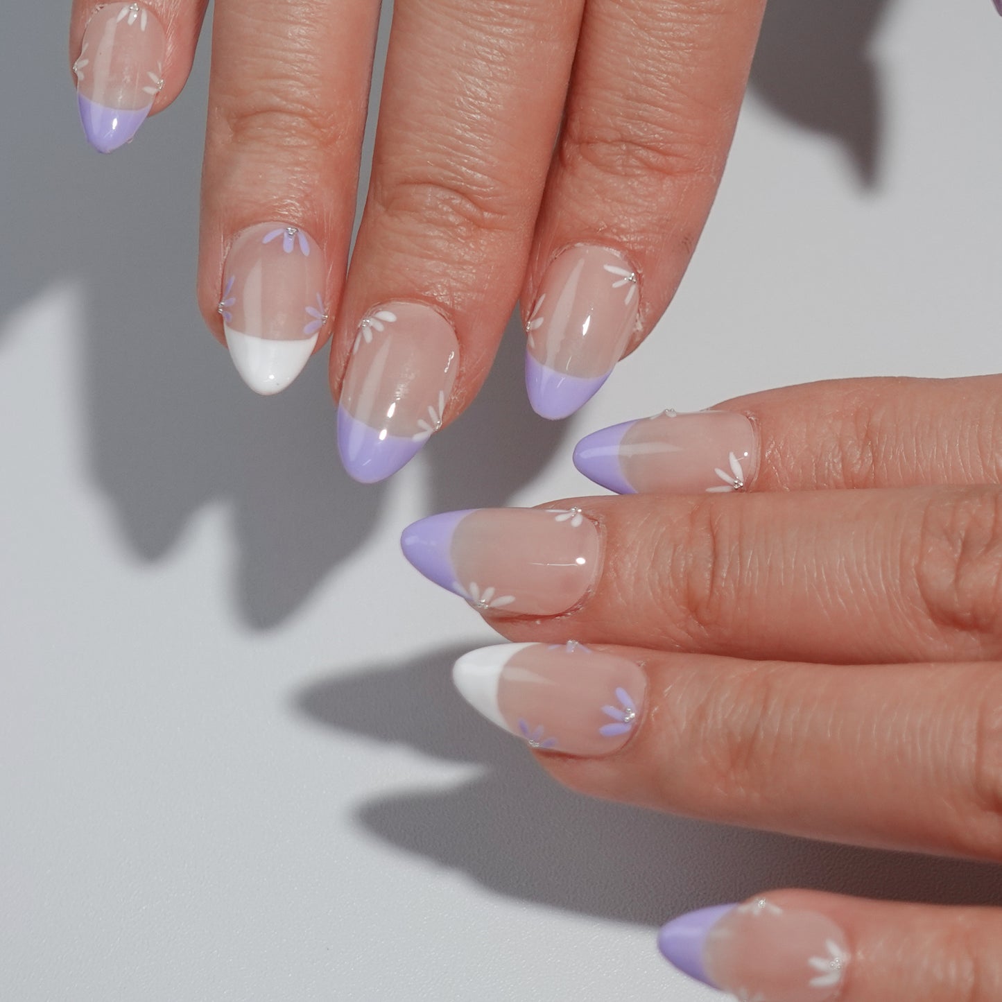 Press-on nails with a nude base, purple and white French tip design, accented with intricate floral details for a refined and spring-inspired look.