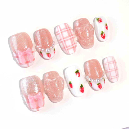 A fruity and feminine press-on nail design featuring pink glitter, plaid patterns, and tiny red strawberries with green leaves. Some nails are adorned with pearls and a 3D bow, creating a playful yet elegant look.