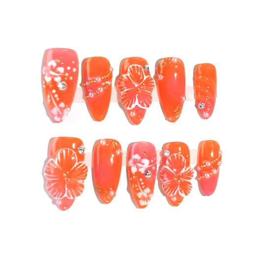 A vibrant coral press-on nail set featuring hand-painted white hibiscus flowers, sparkling rhinestones, and golden accents, evoking the warmth and beauty of a tropical getaway. Perfect for summer-inspired looks.