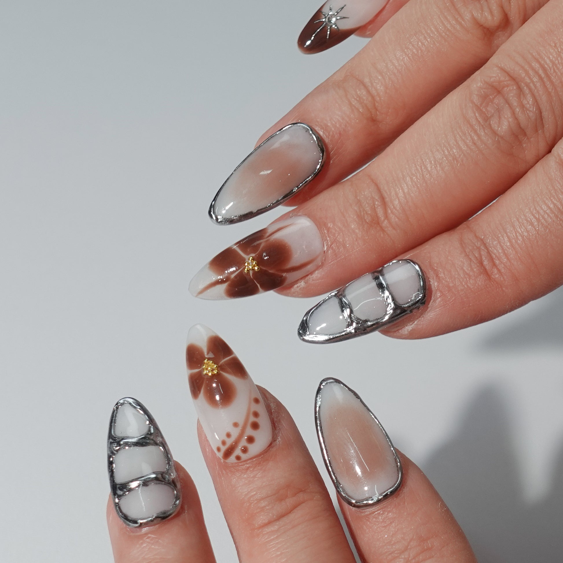 Press - on nails in chocolate brown, with metallic accents, geometric and floral patterns.