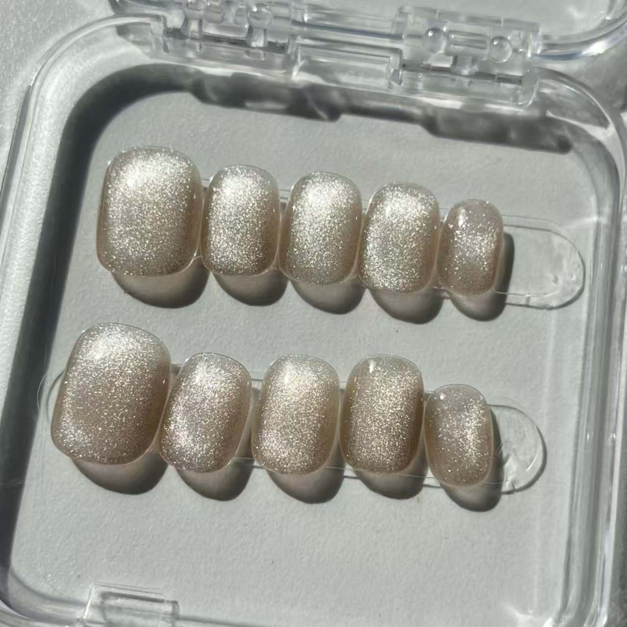 A set of champagne - colored glittery press - on nails neatly arranged in a transparent case. The nails have a sparkling finish, ideal for adding a festive and luxurious touch to your look, perfect for weddings or parties.