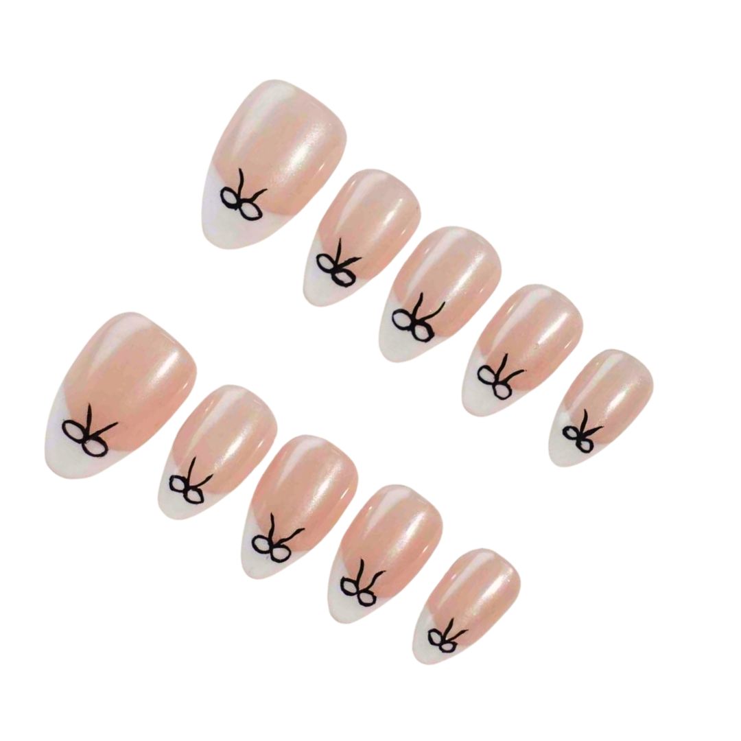 Press-on nails featuring a glimmering nude base with white tips and delicate black bow designs, perfect for a chic and elegant look.